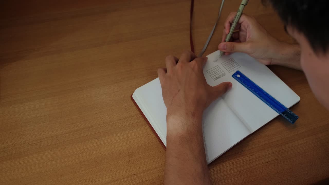 Static shot of male student writing lines for a calendar view inside a bullet journal over wooden desk