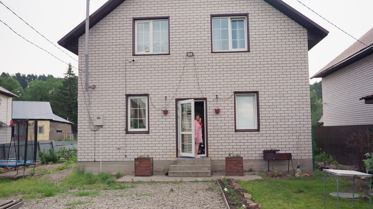 Woman In Pink Stands Outside, Female Figure In Pink Appears Motionless, Lady Dressed In Pink Pauses Quietly At Doorway, Woman Wearing Pink Gown Is Standing Calmly At Open Door Of Suburban House