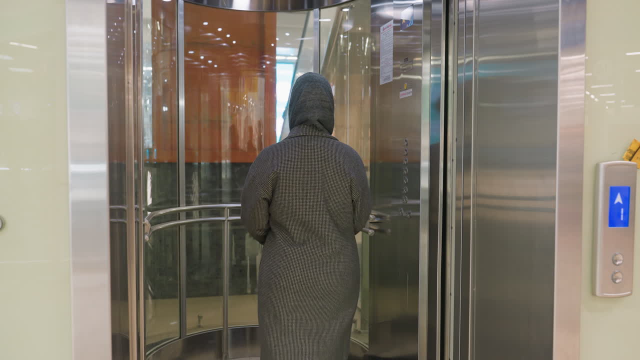 woman in stylish coat enters glass elevator from hallway with surrounding reflections and interior lights seen in transparent enclosure inside modern shopping mall environment