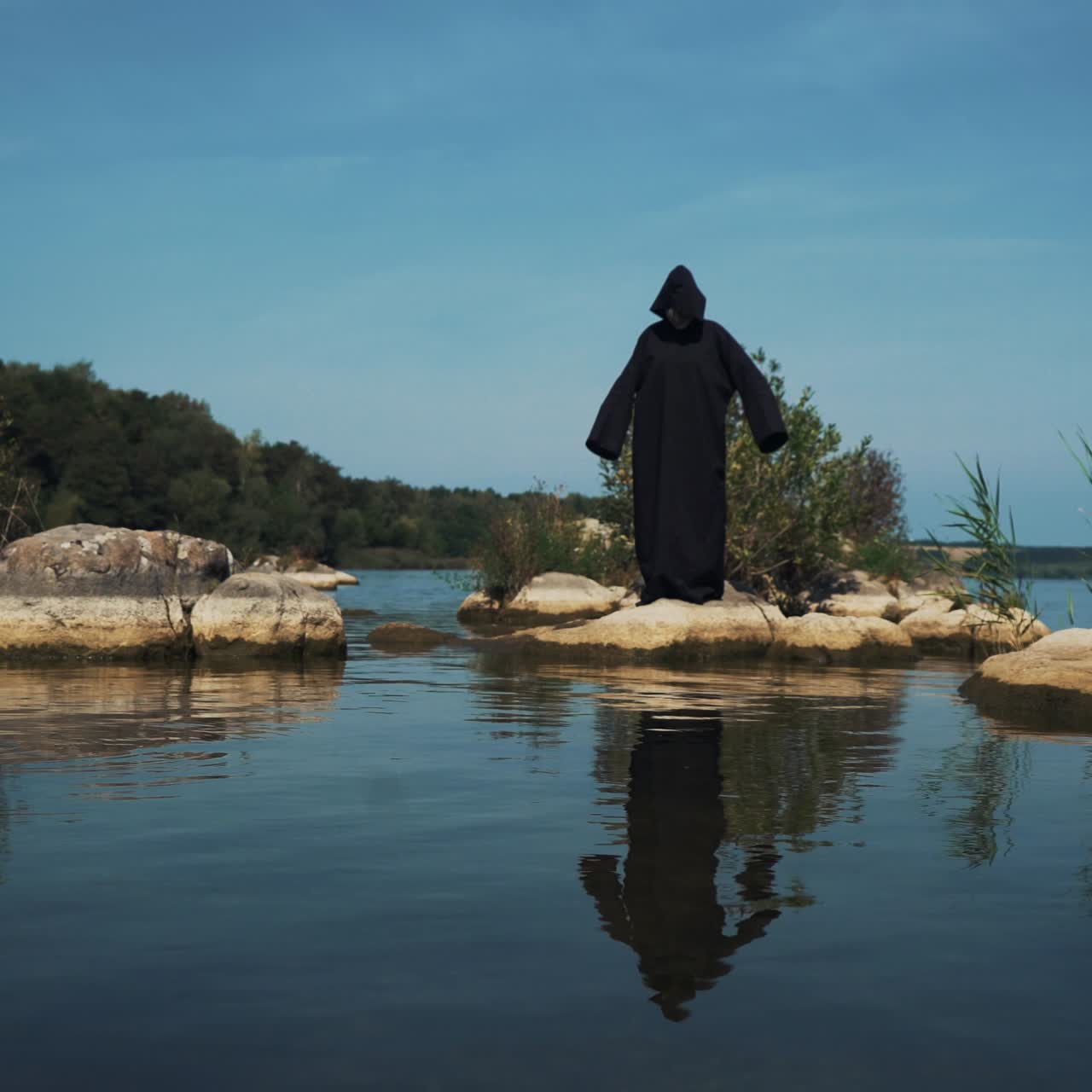 Evil witch in black cloak with hood on stones near the river. Divination by the water. Halloween costume.