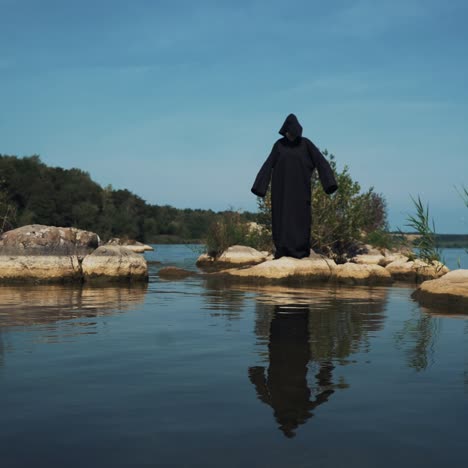 evil witch in black cloak with hood on stones near the river. divination by the water. halloween costume.