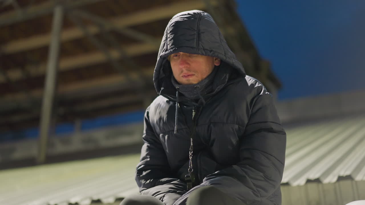 검은 옷과 후드 재을 입은 남자가 다리 사이에 손을 고 혼자 앉아 있으며, 그는 희미하게 밝은 배경으로 경기장에 있습니다.