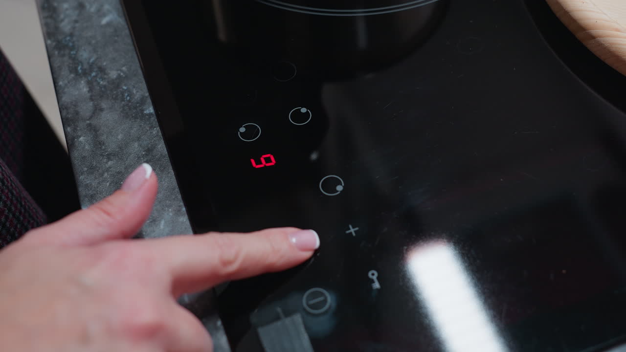 primer plano de la mano del cocinero ajustando los ajustes de temperatura en la estufa eléctrica moderna, el panel de control táctil refleja la mano del cociner y la cocina circundante mientras preparan una comida