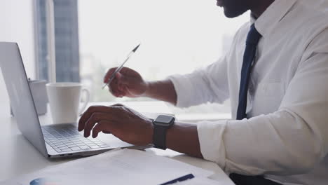 Close-Up-Of-Businessman-Working-At-Desk-On-Laptop-In-Modern-Office-Checking-Data-On-Smart-Watch