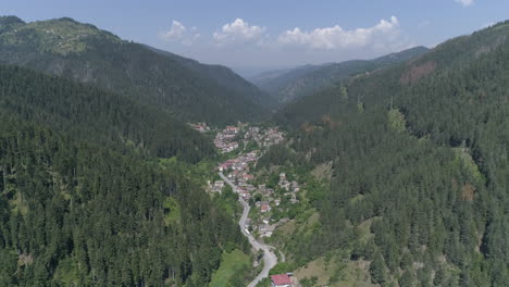 Aérea:-Volando-Sobre-Un-Pequeño-Pueblo-En-Bulgaria-Ubicado-Entre-Dos-Colinas,-Rodeado-De-Bosques-De-Coníferas