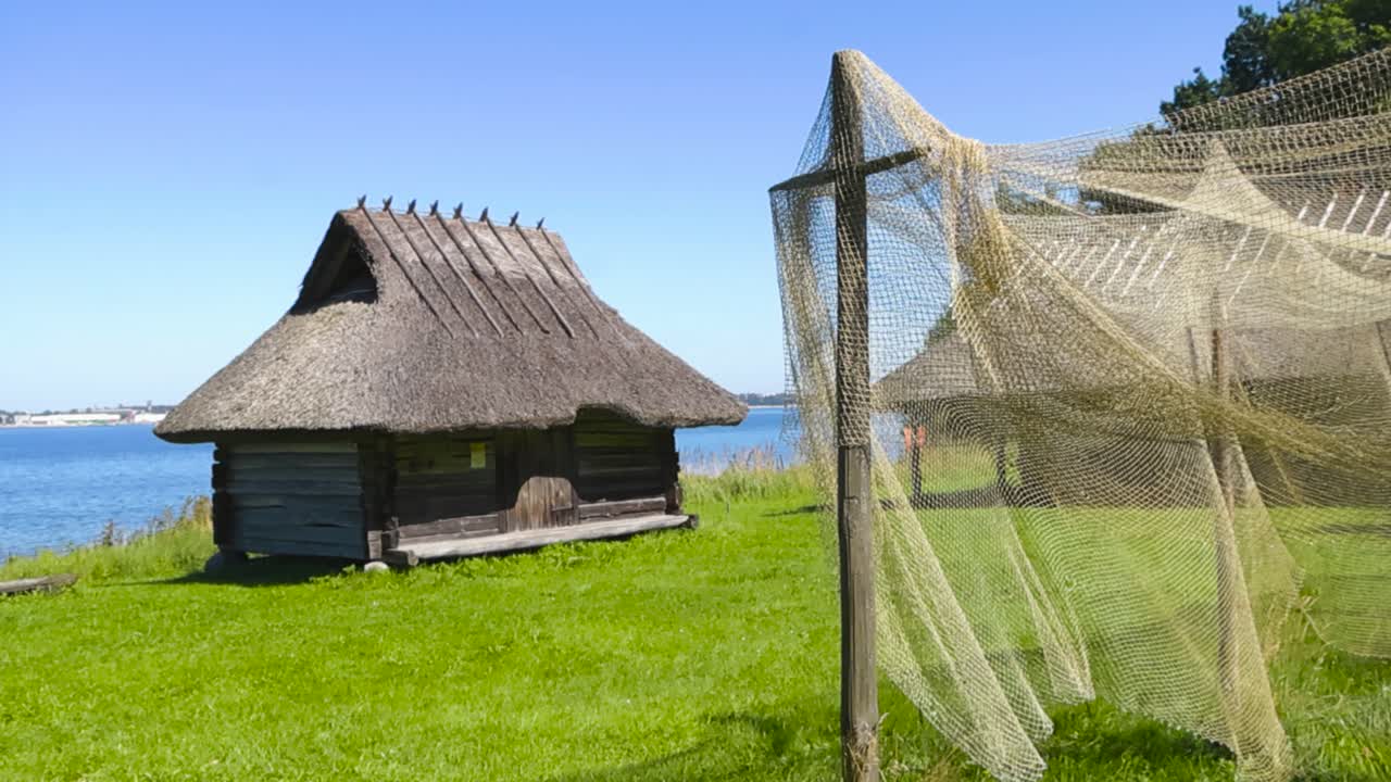 Old yellow and traditional vintage fishing nets drying in the wind in front of old log fishing village cabins with thatched roofs at baltic sea sunny shoreline during summer day. Green grass in garden