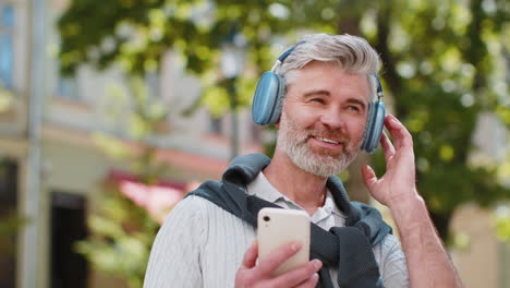 happy mature man in wireless headphones choosing listening music in smartphone application outdoors