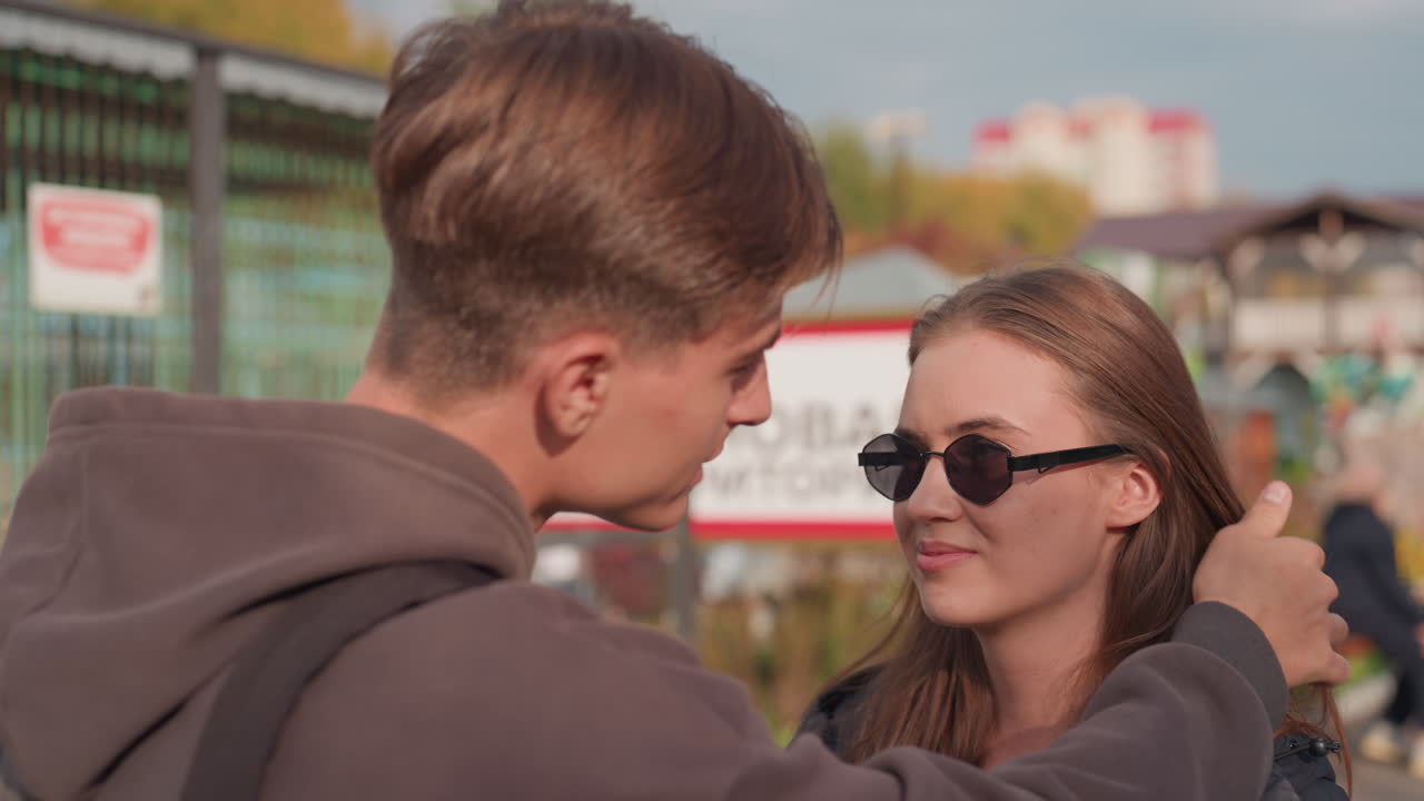 Hvit tenåring justerer forsiktig håret og setter på solbriller, gutt viser omsorg på et utendørs marked, nært og varmt høstlys, uttrykk for tillit og hengivenhet, ærlig filmatisk stemning
