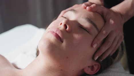 close up of woman with eyes closed lying on towel-covered table as therapist performs gentle forehead massage, surrounded by calm environment with soft light and gray curtain