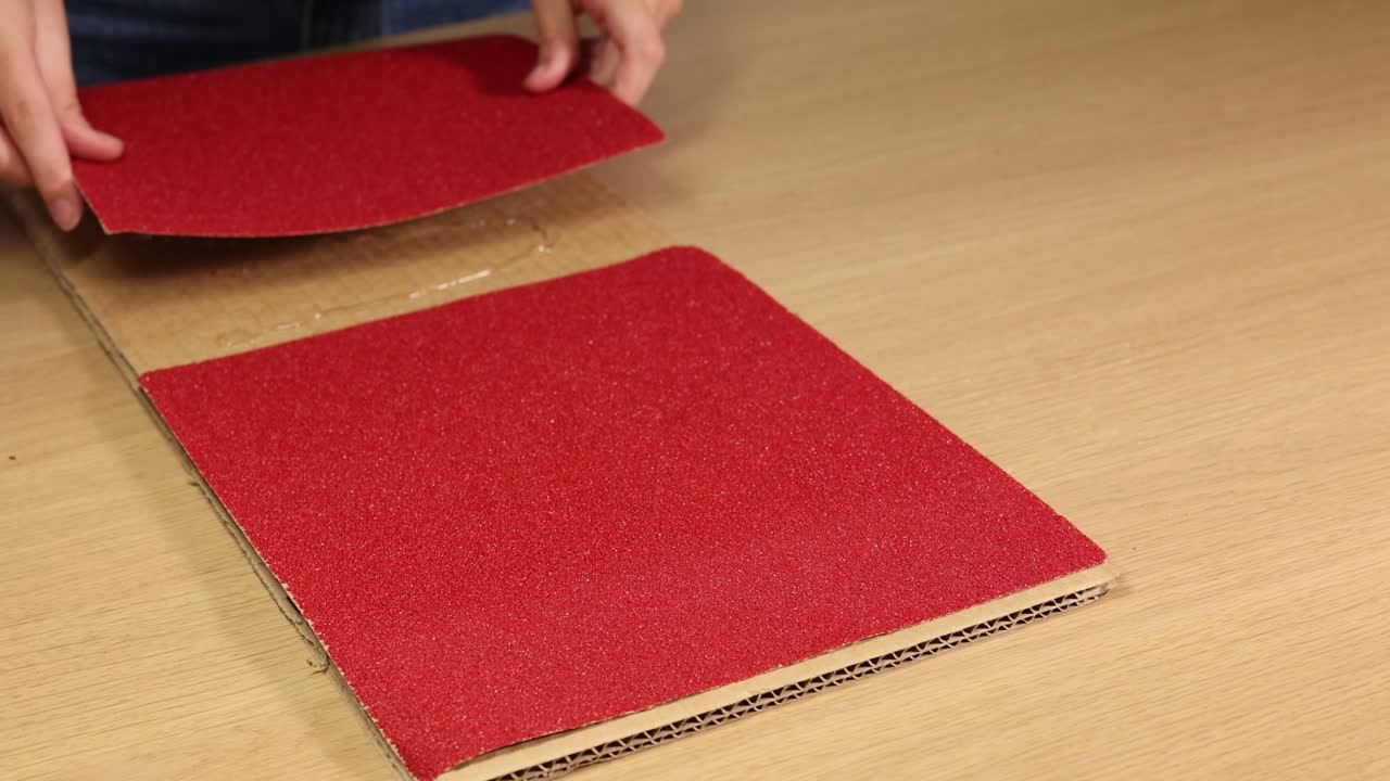 Hands carefully place red sandpaper sheets on a cardboard surface, demonstrating a precise and methodical arrangement technique