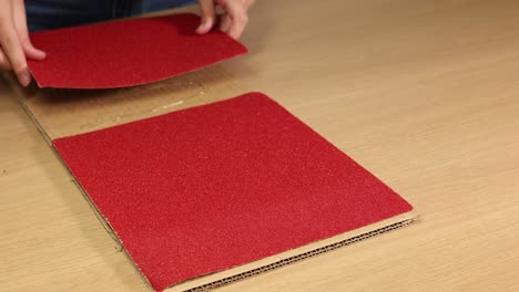 hands carefully place red sandpaper sheets on a cardboard surface, demonstrating a precise and methodical arrangement technique