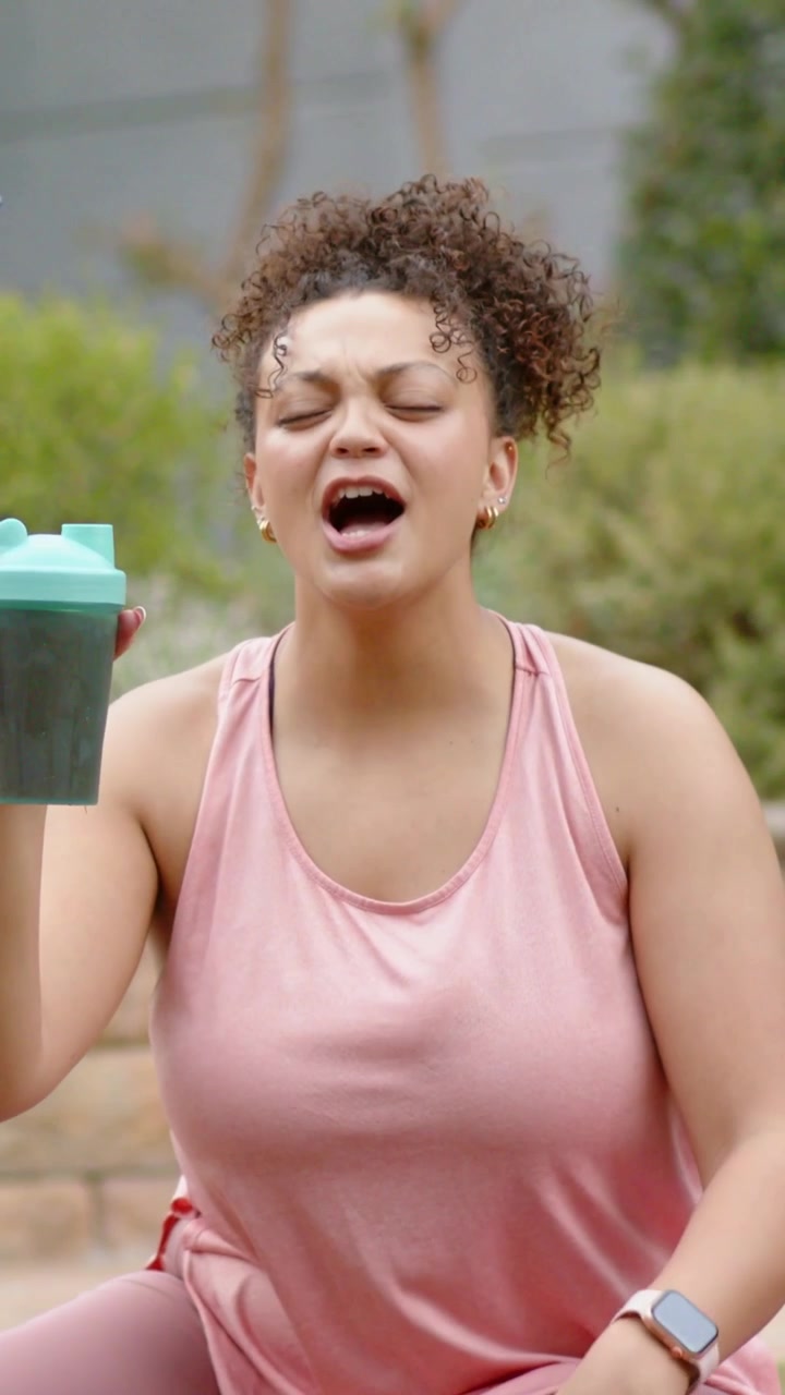 Video verticale: Donna in completo da allenamento rosa sorseggia, alza uno shaker color verde acqua sul patio, assaggiando una tazza offerta.