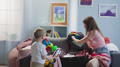 boy carries toys mother puts clothes into suitcase at home