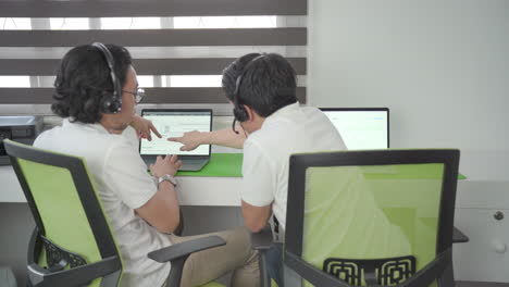coworker helping employee in headsets with new software, pointing at monitor and speaking, back shot