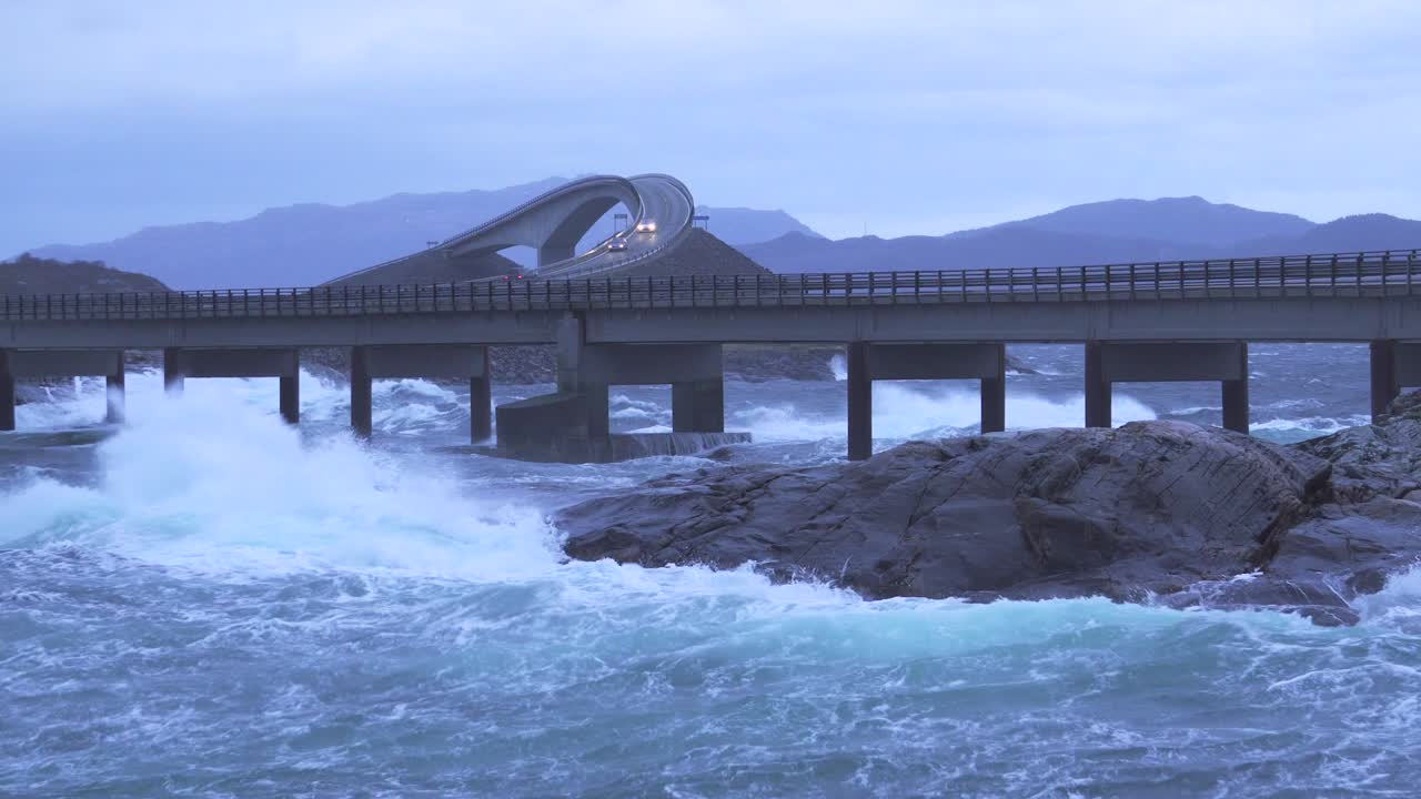 Waves crashing upon rocks next to a bridge, slight zoom in.