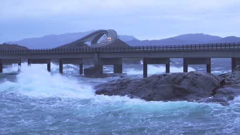 waves crashing upon rocks next to a bridge, slight zoom in.
