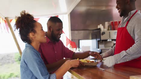 Sonriente-Dueño-De-Un-Camión-De-Comida-Afroamericano-Sirviendo-Comida-A-Una-Feliz-Pareja-Diversa