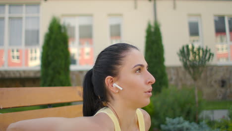 a young woman with headphones performs push-ups on a bench in a city park in slow motion. training a young woman on a bench against the backdrop of the city and houses