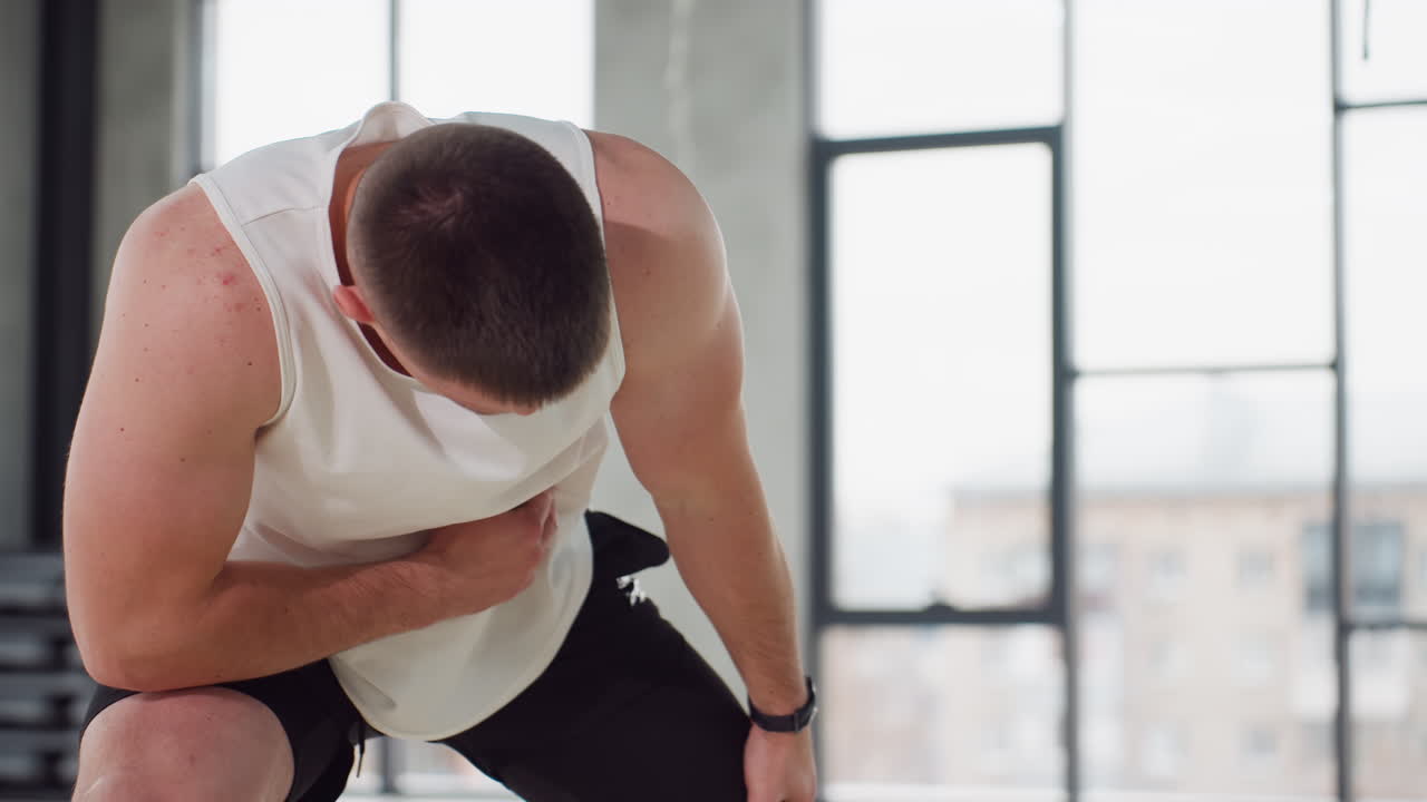 Close up of gym bro performing side lunge warm stretch in bright modern gym wearing white tank and black shorts balancing on light wooden floor under large windows with hanging straps overhead
