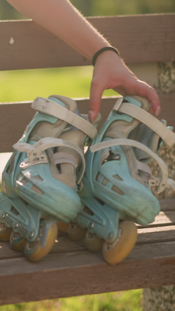 Young white lady drops cyan roller skates on wooden bench and sits beside it with abdomen slightly exposed, sunlight reflects on her with greenery background