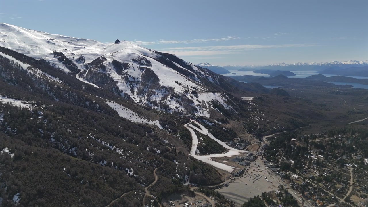 Vista aerea del Cerro Catedral in Patagonia, con cime innevate, sentieri tortuosi, fitte foreste e laghi distanti sotto un cielo azzurro e limpido, che cattura una bellezza serena, ripresa orbitale con drone.
