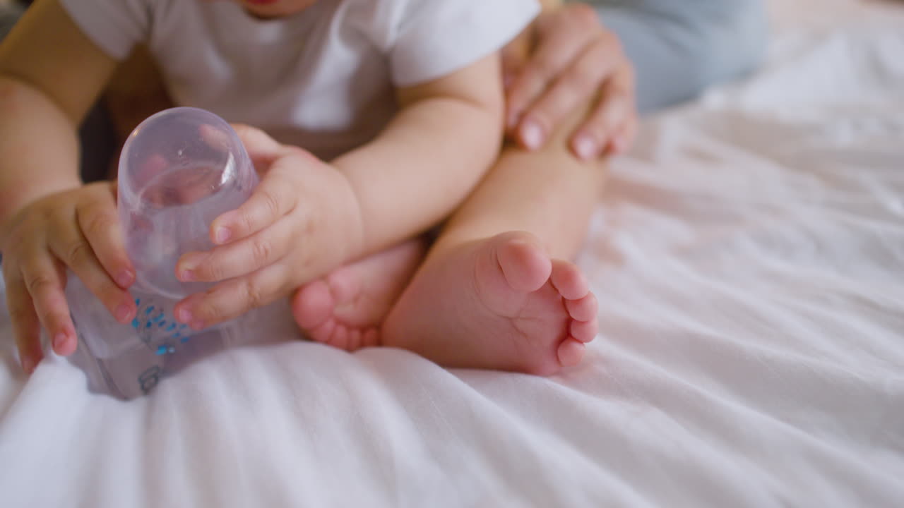 아기가 젖병을 가지고 놀고 엄마가 침대에서 아기의 다리를 애무하는 동안 카메라는 아기의 발에 초점을 맞춥니다.