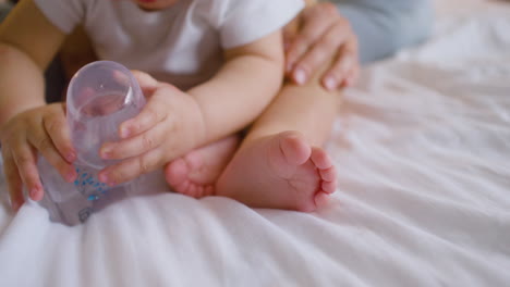 la cámara enfoca los pies de un bebé mientras el bebé juega con el biberón y su madre le acaricia las piernas en la cama