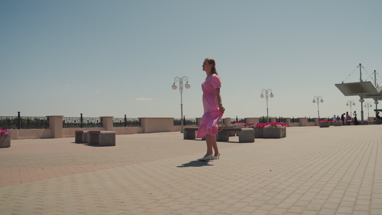 Donna in abito rosa che passeggia sul lungomare lungo un viale costiero illuminato dal sole, con la brezza che le solleva la gonna, lampioni e fioriere che costeggiano l'ampia spianata pavimentata, panchine vuote e orizzonte marino in lontananza, in tutta tranquillità