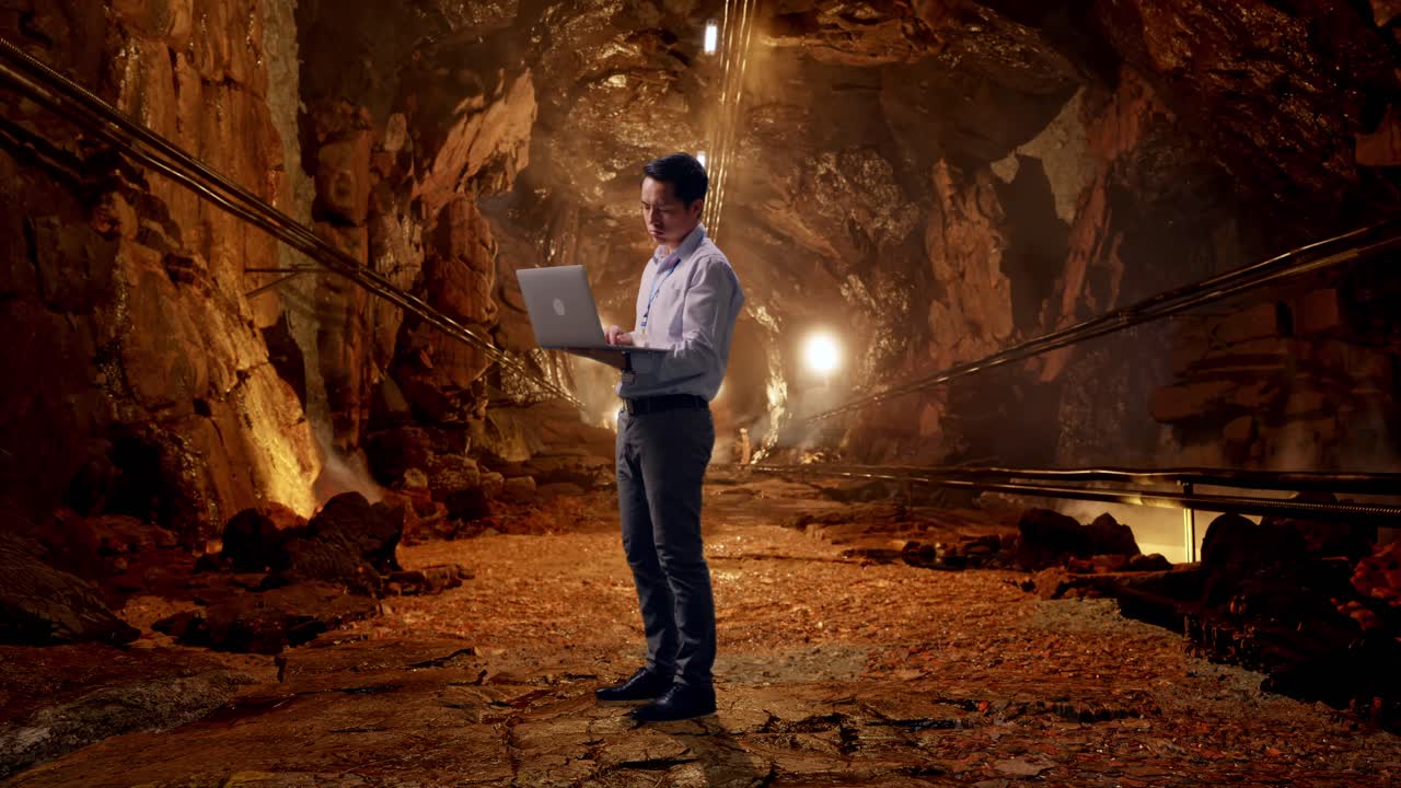 Full Body Side View Of An Asian Male Professional Worker Standing With His Laptop In Underground Mine Tunnel, Typing And Dissapionted With Nodding His Head