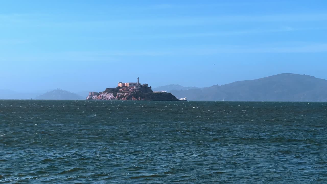 Vista lejana de la prisión de la Isla de Alcatraz en la Bahía de San Francisco. Histórica prisión insular rodeada de aguas agitadas de color azul oscuro bajo un cielo soleado y despejado en California.