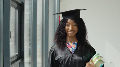 une jeune femme afro-américaine diplômée d'une université se tient avec des manuels à la main près d'une grande fenêtre moderne avec des cadres noirs.