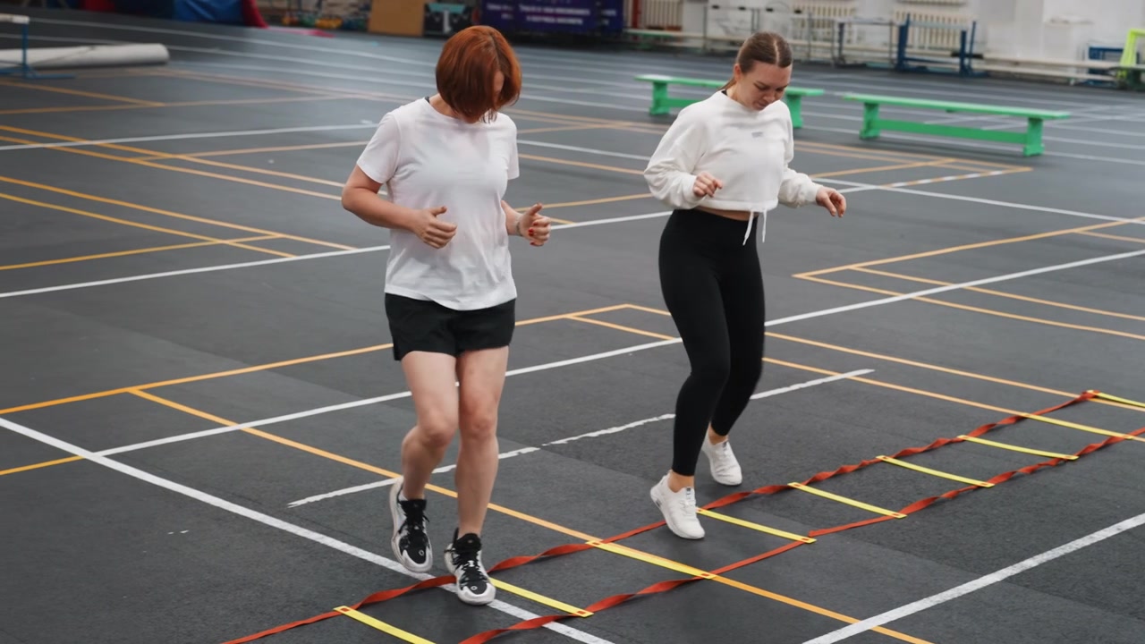 Sidebyside ladder footwork practice partners coordinate steps and rhythm on marked court, cooperative tempo, light banter, repetition for timing and balance, indoor training hall