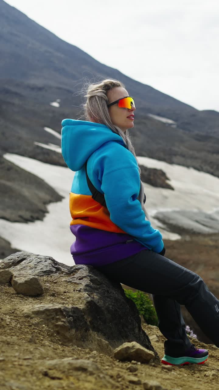 Ung kvinde i farverig hættetrøje sidder på en sten i et bjerglandskab.