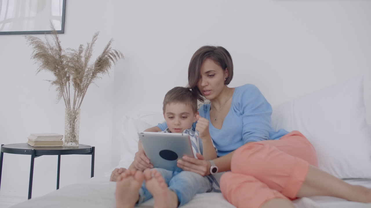 en el dormitorio blanco madre e hijo miran la pantalla de la tableta y se ríen. familia feliz en la cama por la mañana leyendo un libro y haciendo clic en el touchpad de la computadora.