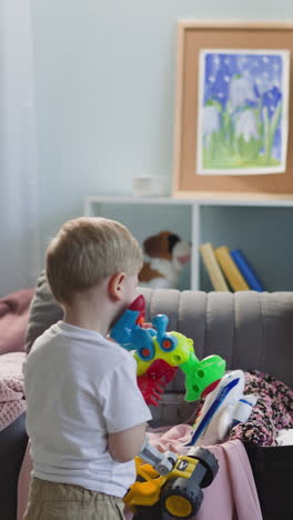 little blond boy carries pile of toys while mother puts clothes into suitcase in living room. preoccupied family prepares for vacation journey