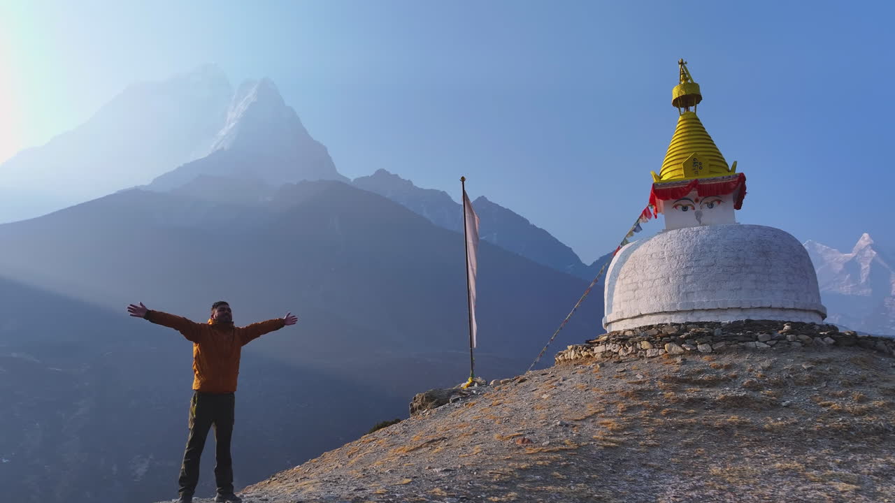 Wide aerial view of a tourist at a Buddhist stupa in Dingboche, Khumbu, Nepal. Mt. Ama Dablam rises in backdrop with mountain reflections on horizon, creating a peaceful, magical Himalayan scene