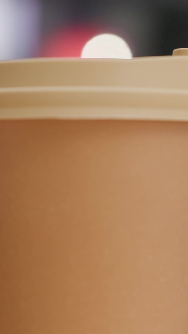 Mano che tiene un contenitore marrone per bevande con coperchio. Posizionato davanti a un'area che mostra una luminosità diffusa. L'oggetto principale mantiene una posizione fissa in mezzo a un'illuminazione sfocata.