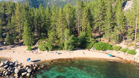 vista aérea de drones de la playa pública en el lago tahoe, california con gente caminando, espeso bosque de pinos en el fondo, aguas azules claras y grandes rocas rocosas