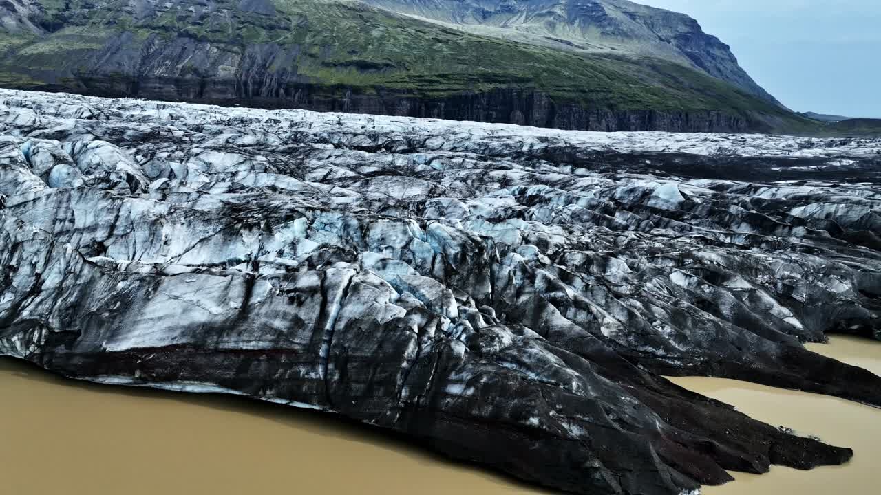 A drone sweeps along the edge of Vatnajökull, revealing dark, cracked glacier ice above milky meltwater with steep volcanic mountains rising in the background