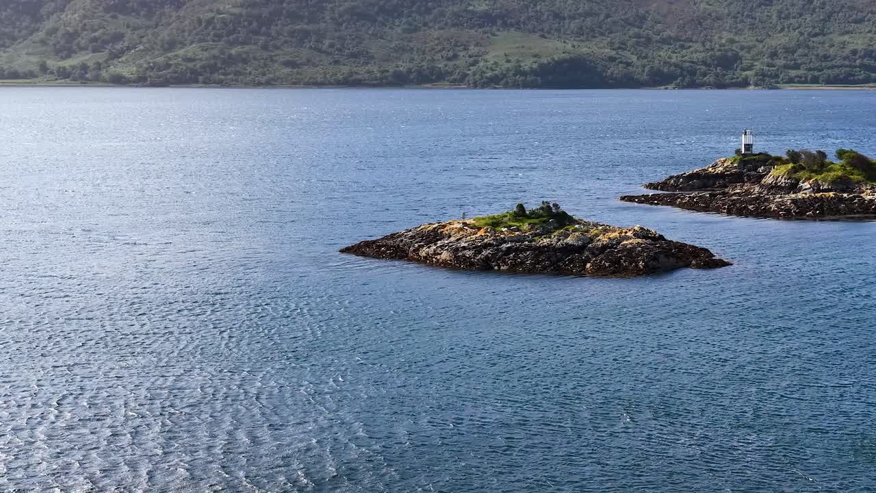 Drone glides over tranquil blue water toward rocky island with lighthouse, lush greenery, natural daylight
