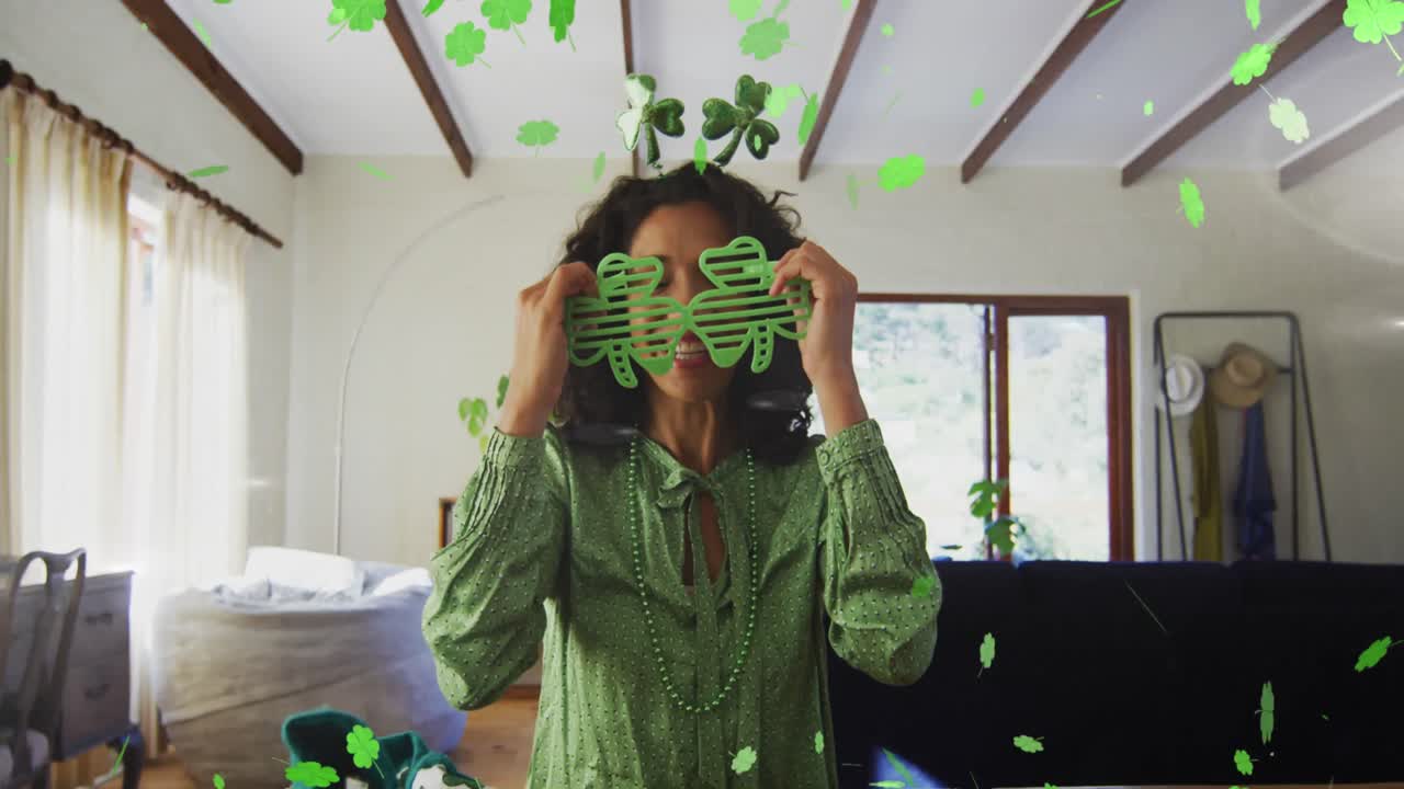 Animation of shamrocks falling and biracial woman in shamrock glasses celebrating st patrick's day