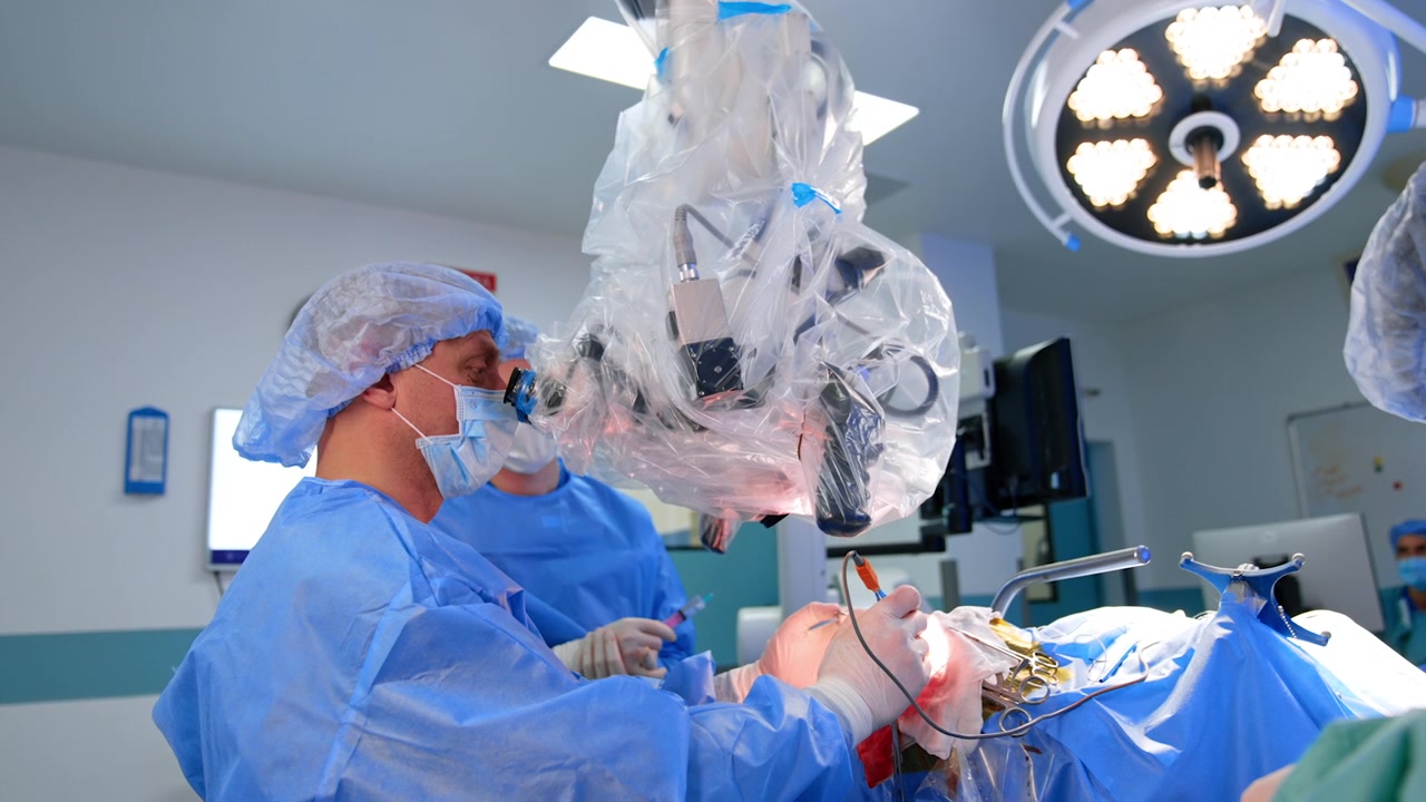 Neurocirujano profesional realiza una operación mirando por el microscopio. Médico asistente sosteniendo una jeringa al fondo.