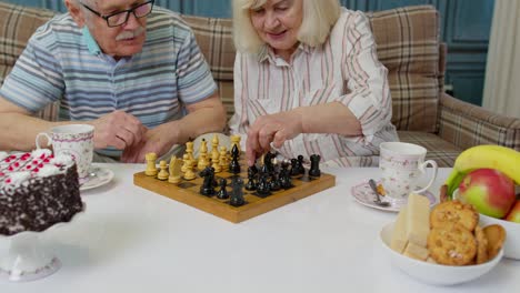 una pareja de ancianos sonrientes abuelo abuela descansando en el sofá bebiendo café, jugando al ajedrez en casa