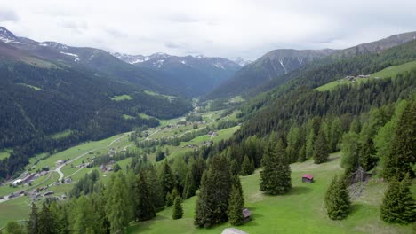 Drohnenaufnahmen-Aus-Der-Luft,-Die-Im-Frühling-Hoch-über-Einer-Blühenden-Almwiese-Fliegen,-Mit-Einer-Schweizer-Alpenblockhütte,-Einem-Wald-Aus-Grünen-Nadelbäumen,-Bergen-Und-Einem-Dorf-Im-Hintergrund