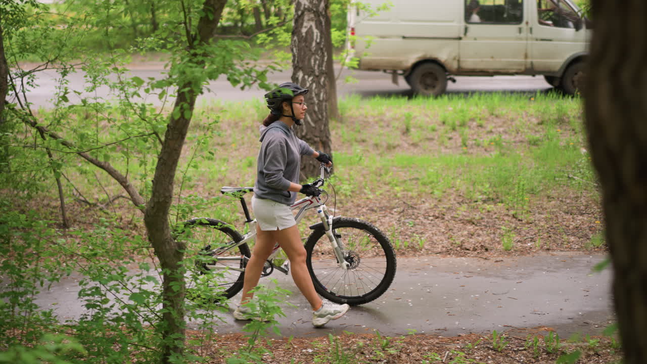 Woman Pushing Bike Through Green Forest Path, Casual Commuter Pause By Trees And Roadside, Helmet On, Shorts And Hoodie, Slow Stroll With Bike Beside, Leafy Branches Framing Scene, Soft Daylight