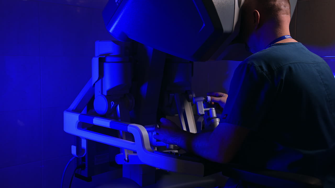 Male doctor in blue uniform sitting his back to camera in front of operational system of Da Vinci robot. Specialist working in dark surgical room.