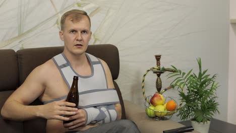 Painful-man-with-a-broken-arm-wearing-arm-brace-sitting-on-a-sofa-drinks-beer