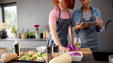pareja feliz diversa en delantales preparando verduras y usando tableta en la cocina, en cámara lenta