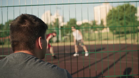 晴れた日に屋外のバスケットボール コートで座ってバスケットボールのトレーニング セッションを見ている若い男の背面図 1