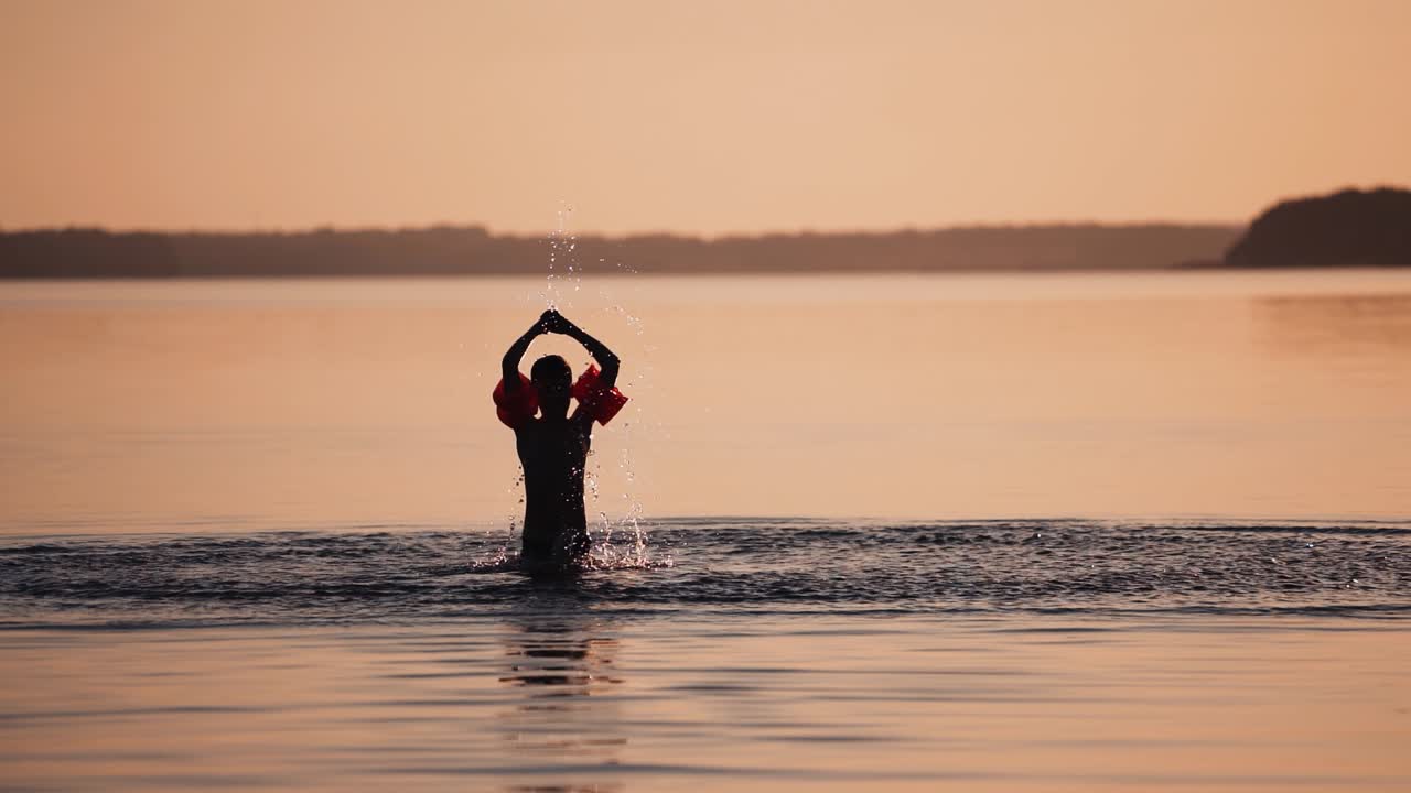 Slow motion of a child in water at sunset. Cute boy having fun in the river alone in the evening. Silhouette of boy splashing water in the lake.